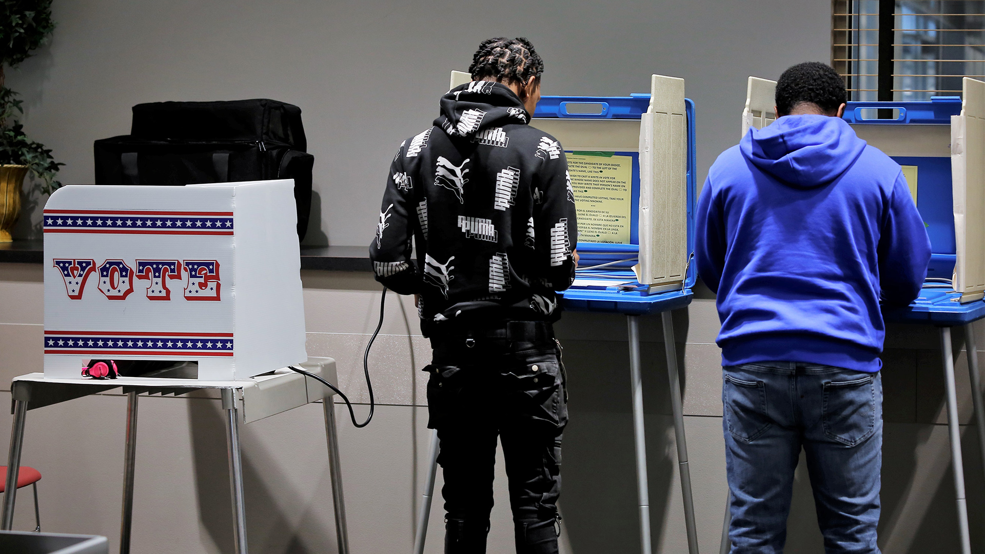 Christophe and Brandon LaSalle stand and face folding voting booths with metal legs and plastic privacy screen walls, in a room with potted plants and a window.