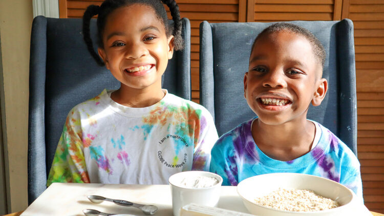 Two children sit at a table, smiling at the camera. At the table in front of them are baking ingredients.