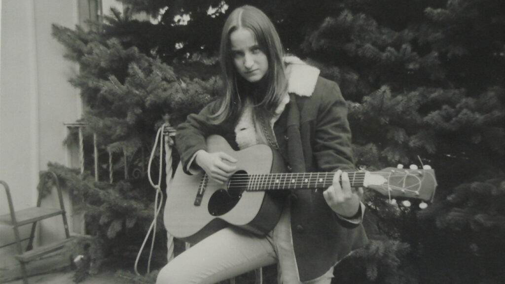 A black and white photo of Lou Sullivan with long hair in the 60s, playing the guitar