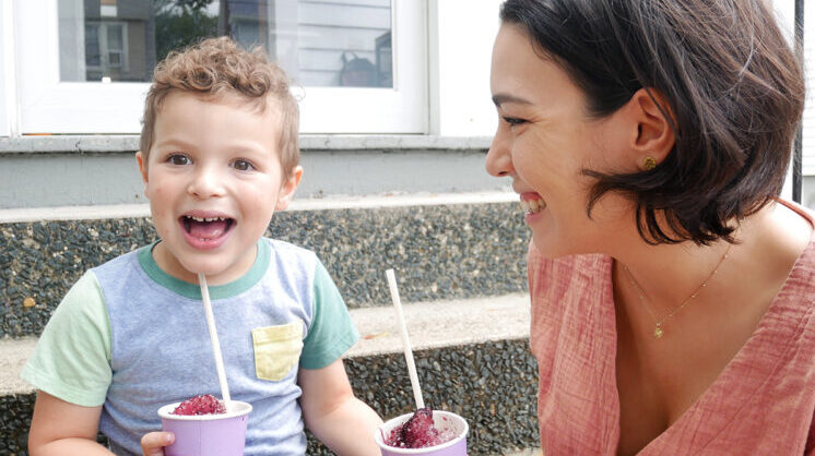 A grown-up and child sit outside together. They're holding cups of piragua, a frozen, fruity treat.