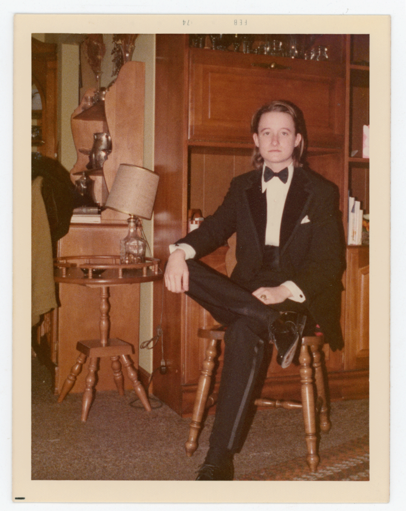 An archival image of Lou Sullivan in a suit, sitting cross legged in a chair.