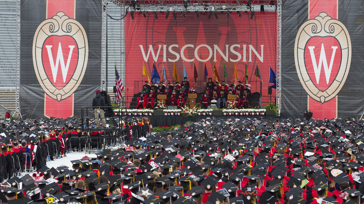 Students wearing graduation attire of caps and gowns are seated in rows facing a stage where faculty and administrators are seated in rises in front of a graphic banner with multiple versions of the UW-Madison W crest and a row of U.S., Wisconsin and other flags.