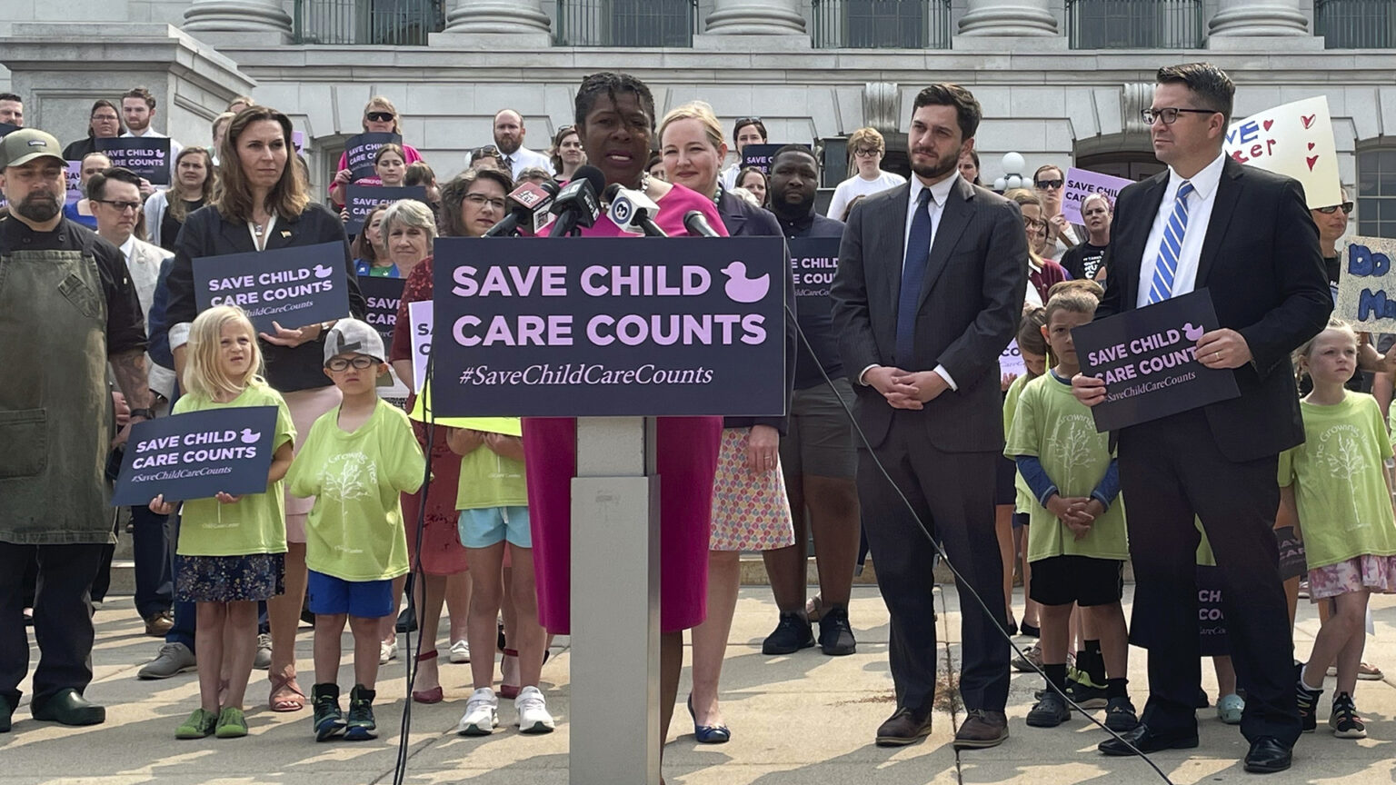 LaTonya Johnson stands and speaks into multiple microphones mounted on a podium affixed with a sign reading Save Child Care Counts, with other adults and children standing to her side and behind, with several holding printed signs with the same slogan and hand-drawn signs, in front of a masonry building.