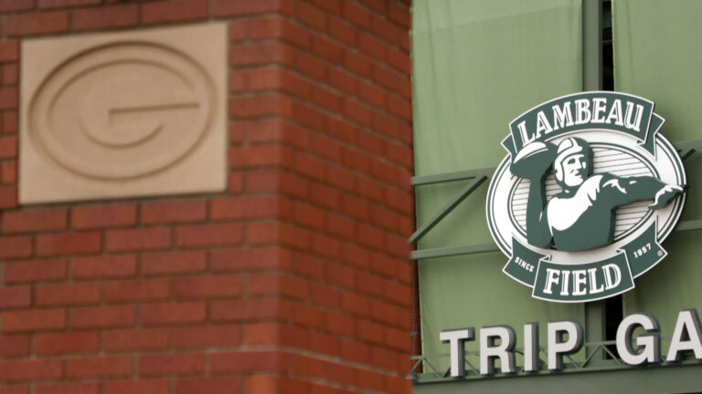 A logo showing a football player throwing a football bracketed by the words Lambeau Field and Since 1957 is stands above a gate entrance to a stadium and next two a brick wall embedded with a concrete Green Bay Packers G logo.