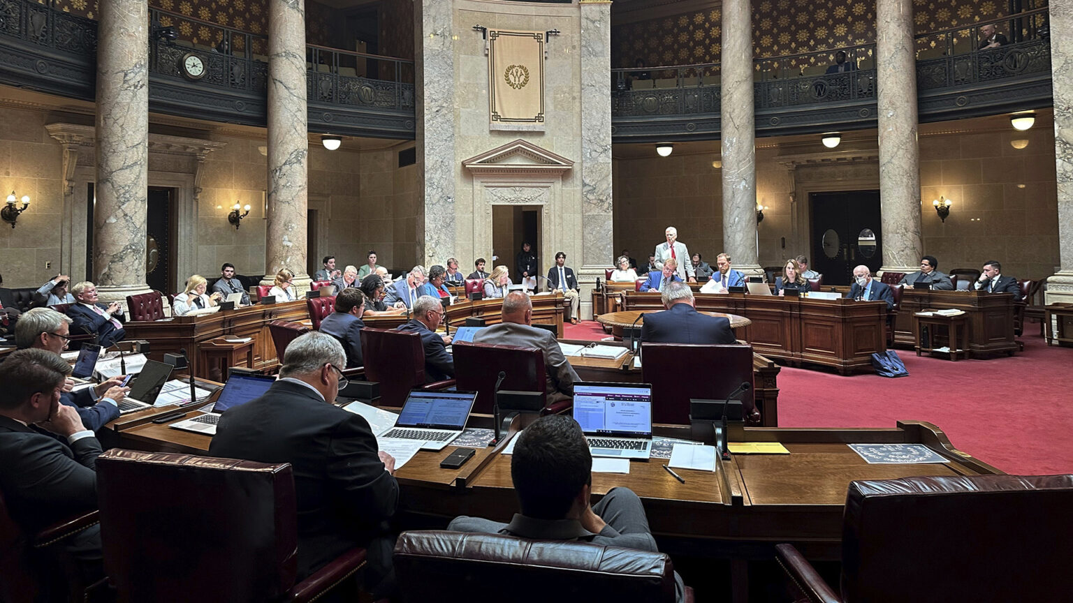 State senators sit at wood desks arranged in a semi-circle and listen to another senator who is standing and speaking, in a room with marble masonry and pillars, and a second-story observation gallery.