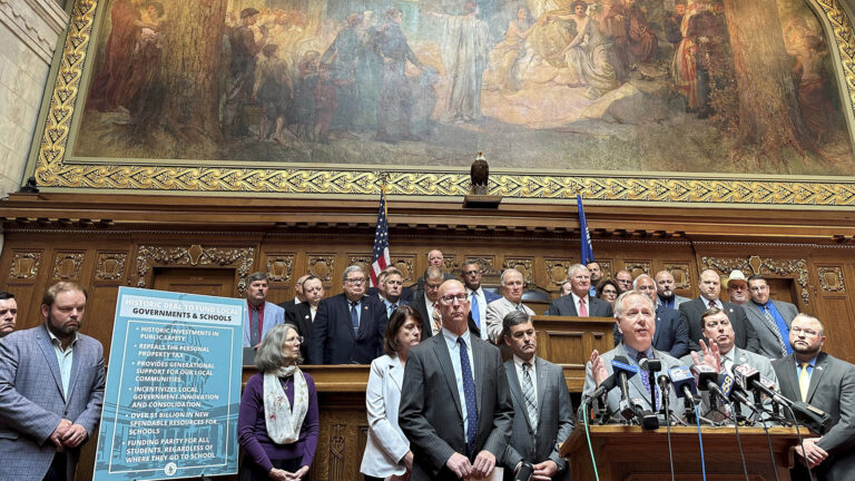 Robin Vos stands behind a podium and speaks while facing more than a half-dozen microphones and gestures with both hands, while other people stand around and behind him along side a poster with the title Historic Deal to Fund Local Governments & Schools and a bullet list, in a room with a large wood dais, wood paneling and moulding, the U.S. and Wisconsin flags, a taxidermy bald eagle and a large painting.