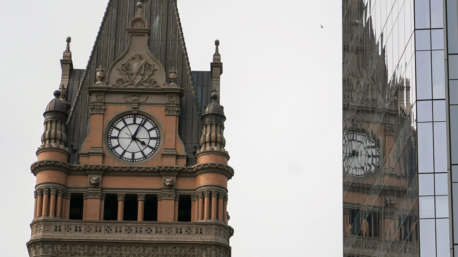 A brick and masonry tower with multiple copper-roofed spires includes a clock face reading 4:04, with half of the structure reflected in the windows of a nearby glass-walled building.