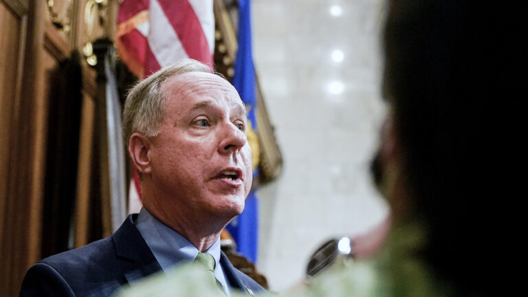 Robin Vos and speaks to reporters while standing in front of U.S. and Wisconsin flags, with a blurry person in the foreground on the right side of the image.