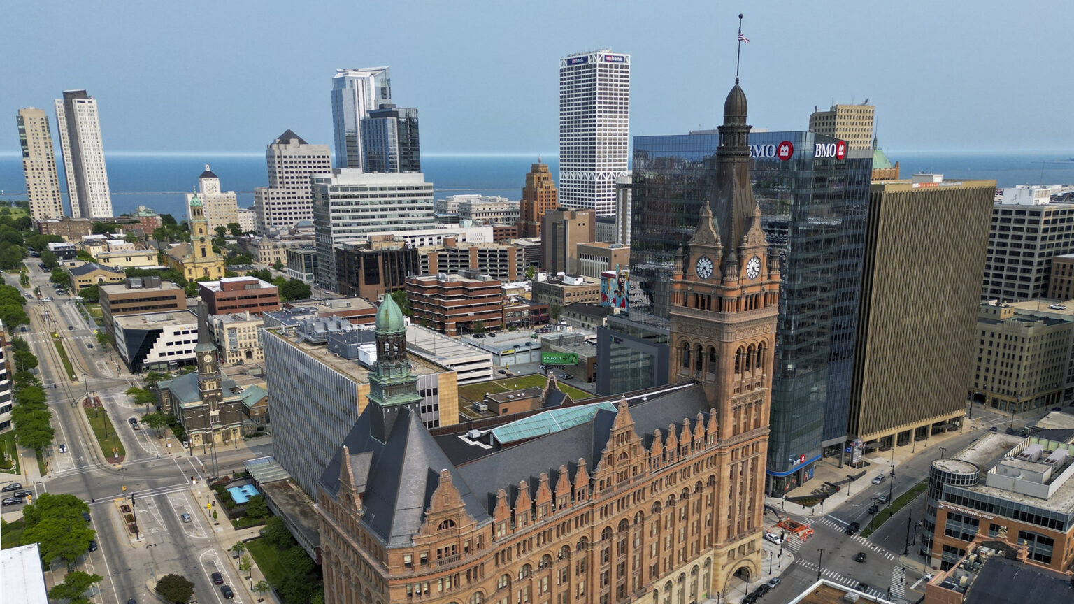 An aerial photo shows a brick and masonry Renaissance Revival style building with multiple spires and a clock tower stands in the foreground of a downtown skyline of buildings of varying heights, with the waters of Lake Michigan visible in the background.