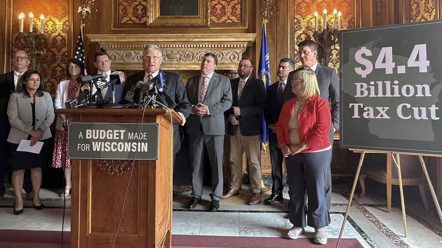 Howard Marklein stands behind wood podium affixed with a sign reading A Budget Made for Wisconsin and speaks into multiple microphones, with other people standing behind him and next to a sign on an easel reading $4.4 Billion Tax Cut in a room with marble flooring, toile wallpaper, brass electric wall sconces, and a fireplace with a carved lintel.