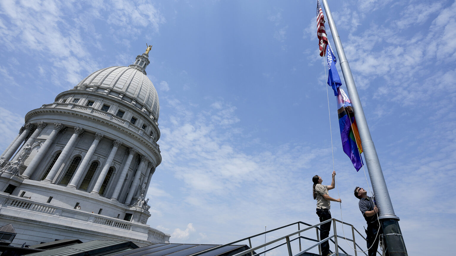 Mindy Fude and Steve Walker stand on a walkway and hold different ends of halyard rope to raise the U.S., Wisconsin and Progress Pride flags on a flagpole, with the skylights of one wing of the Wisconsin State Capitol and its dome in the background.