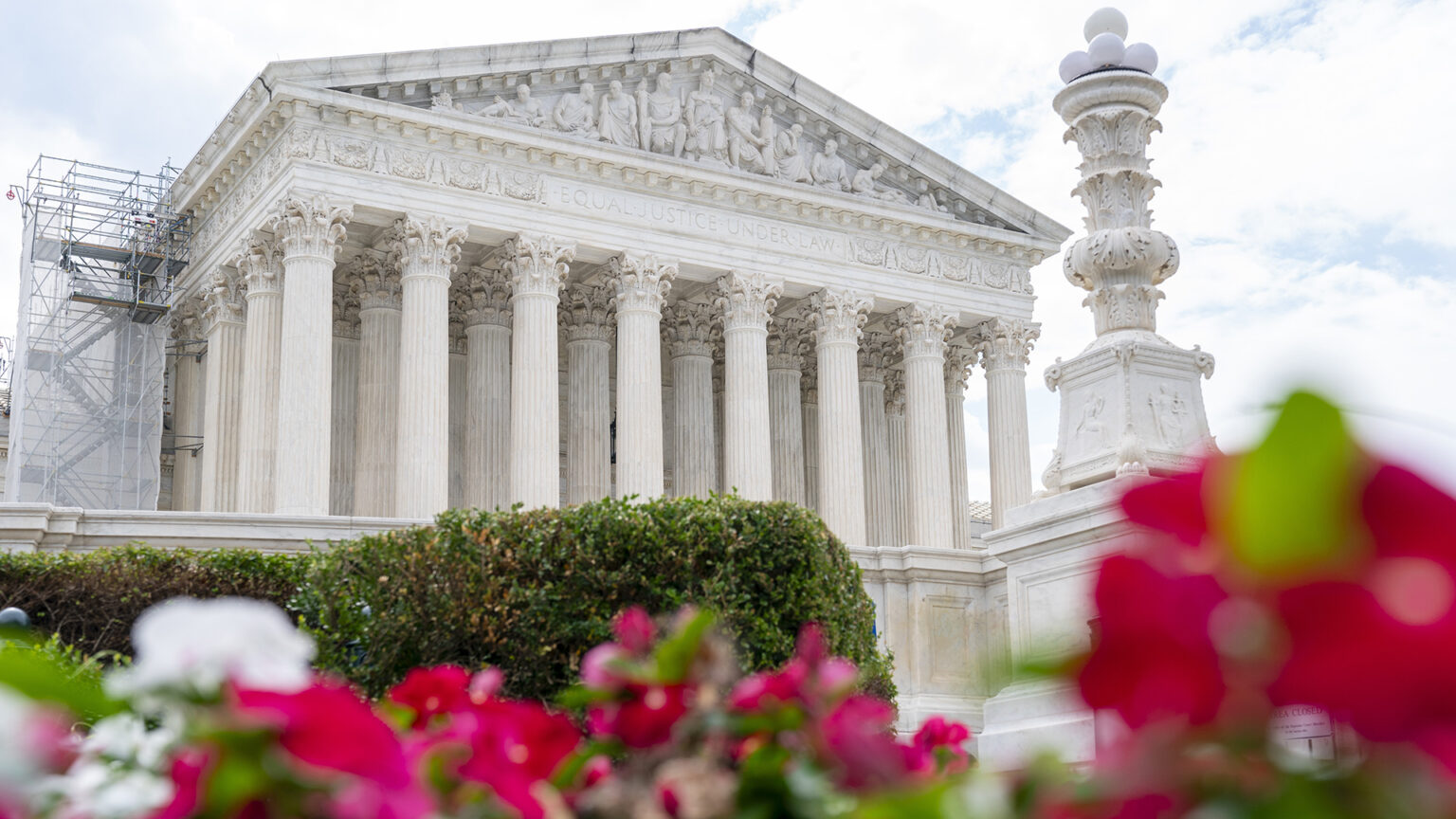 A multi-story scaffold with stairs stands next to the side of a Neoclassical marble building with a double row of columns supporting a pediment with carved words reading Equal Justice Under Law and a frieze with a series of carved figures, with  a pillar topped by a light fixture along with bushes and flowers in the foreground.