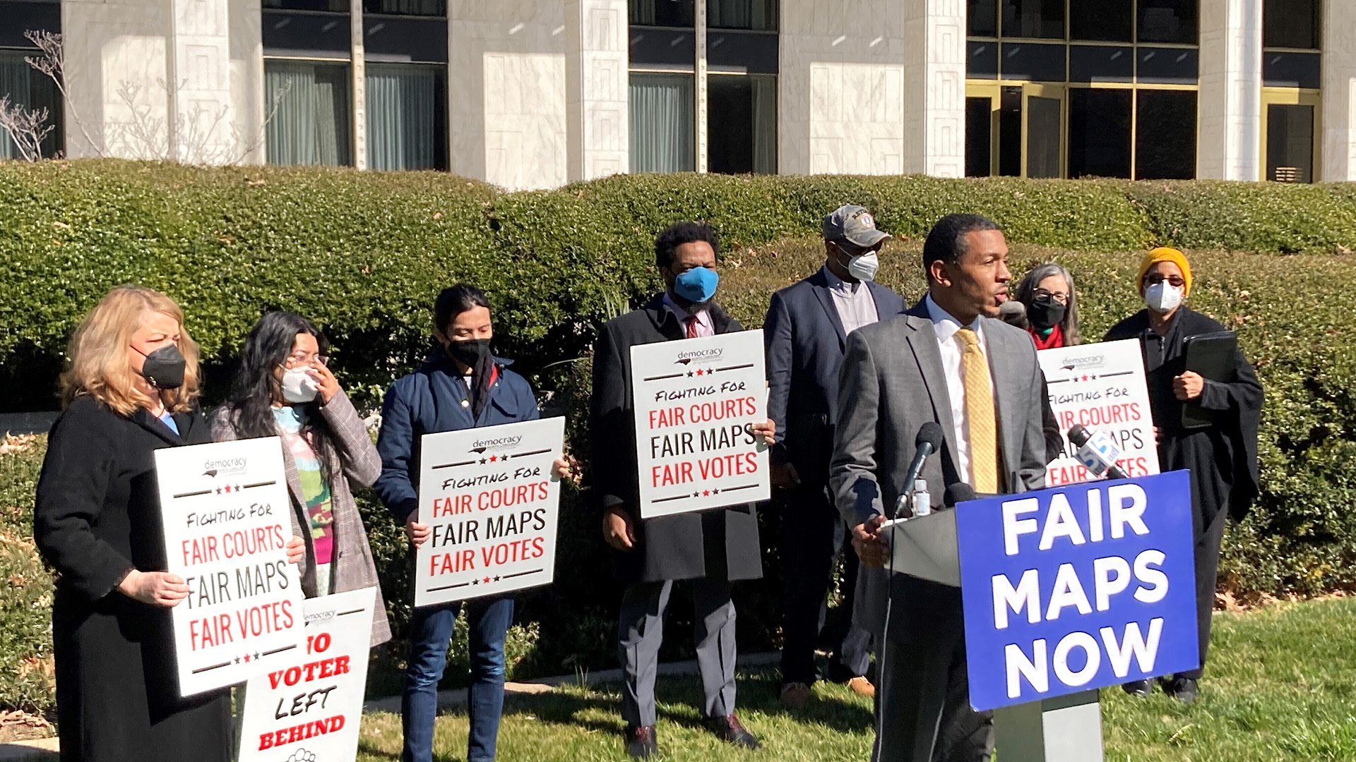 Reggie Weaver speaks while standing at a metal podium with multiple microphones and a sign reading "Fair Maps Now," with multiple other people standing behind him holding printed signs reading "Fighting for Fair Courts, Fair Maps, Fair Votes," with a row of hedges and a building in the background.