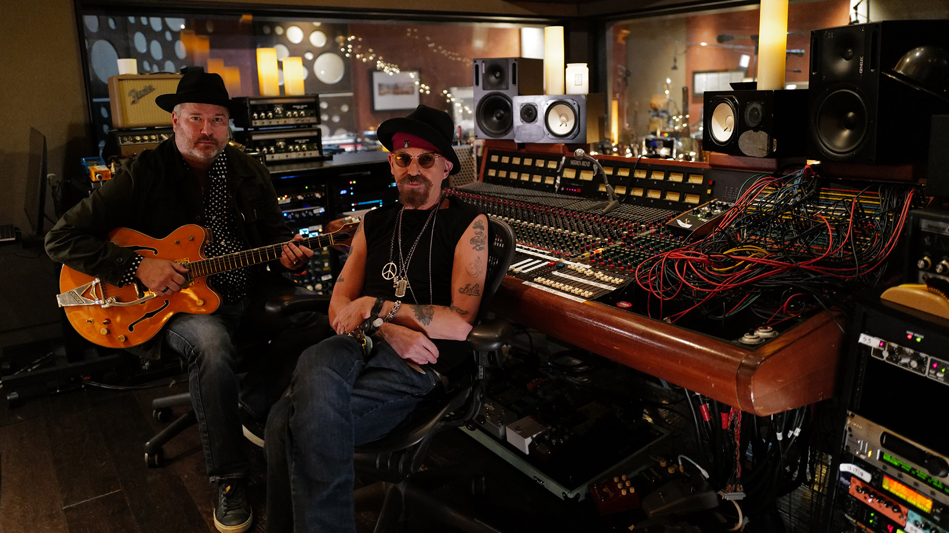 Two men sit in chairs inside a music studio. They smile while facing the camera.