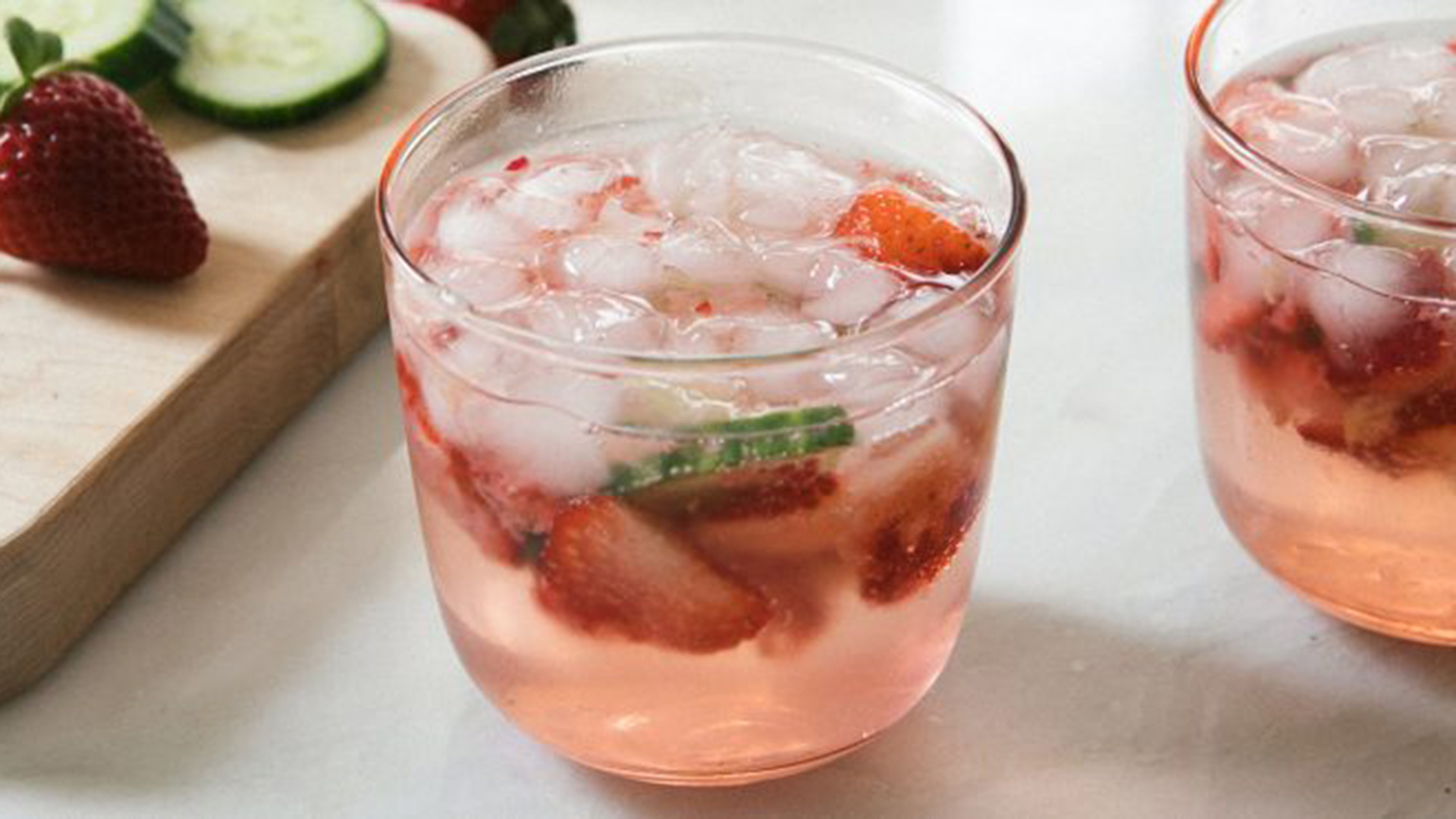 A clear glass cup filled with pink strawberry liquid, ice and sliced strawberries.