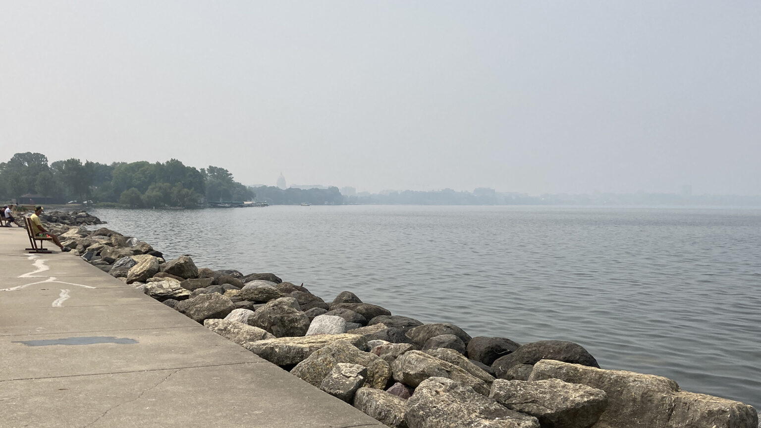 People sit on park benches on a concrete walkway atop a stone breakwater in a lake, with trees and skyline on a receding shoreline obscured by haze and smoke in the air.