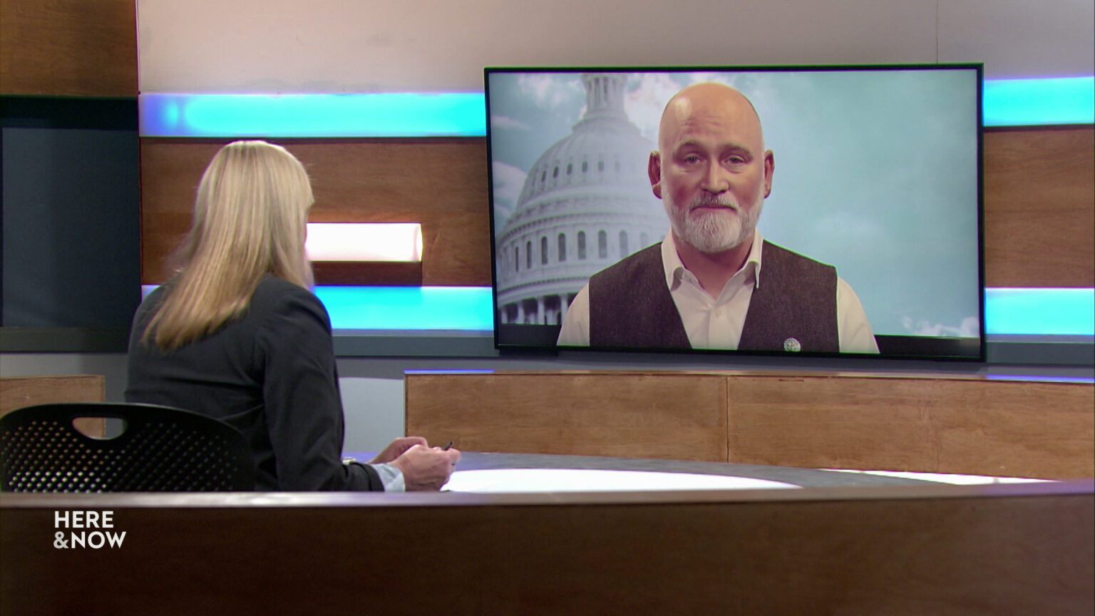 Frederica Freyberg sits at a desk on the Here & Now set and faces a video monitor showing an image of Derrick Van Orden.