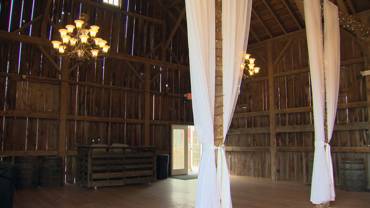 Metal chandeliers and long drapes hang from the rafters of the interior of a barn with wood-slat walls, with multiple barrels and a bar set against the rear wall next to an open door.