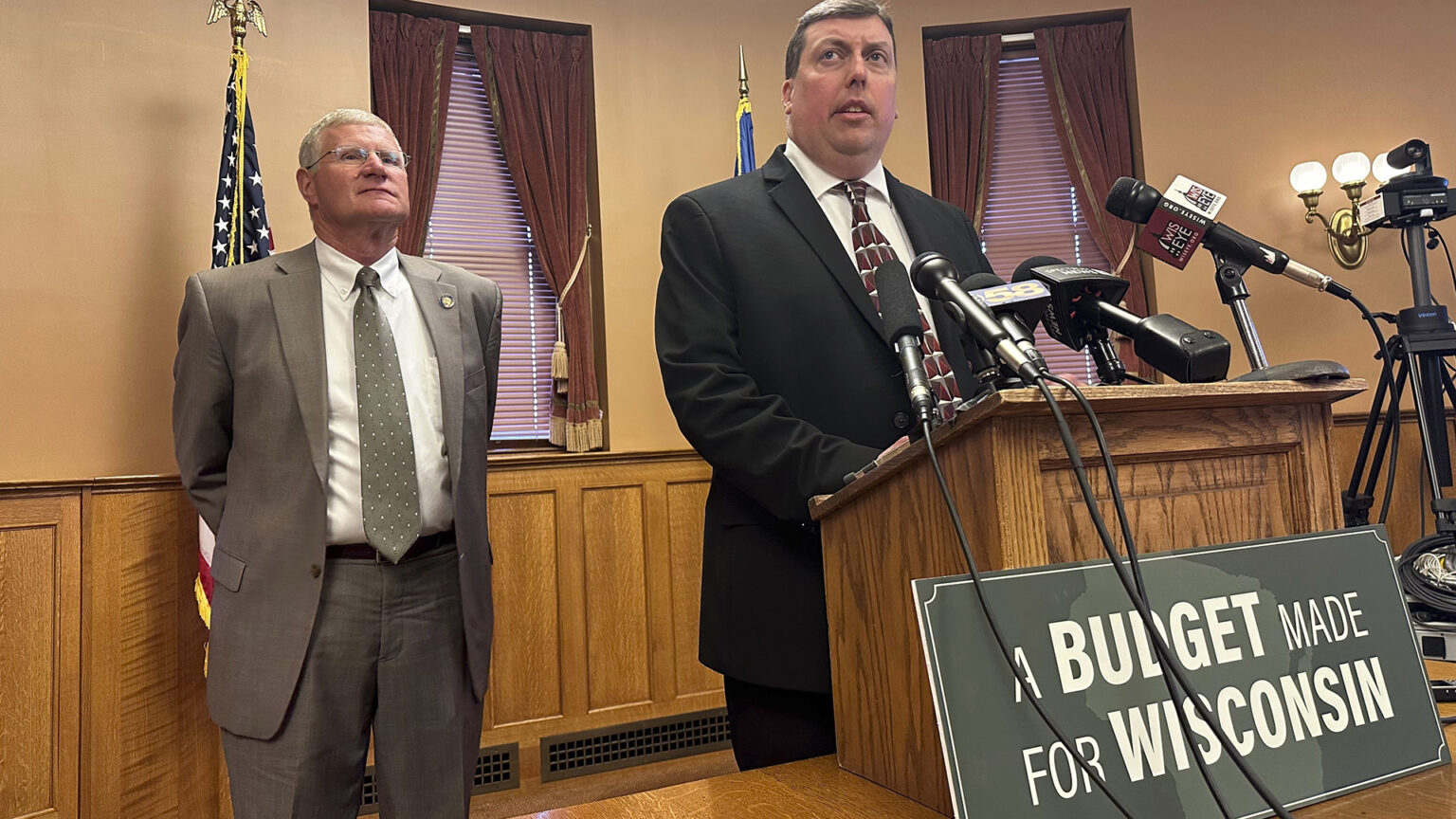 Mark Born speaks into multiple microphones while standing at a table-top podium with a sign at its base reading A Budget Made for Wisconsin, with Howard Marklein standing behind him in a room with the U.S. and Wisconsin flags, a wall sconce lighting fixture, and windows with curtains tied open and closed blinds.