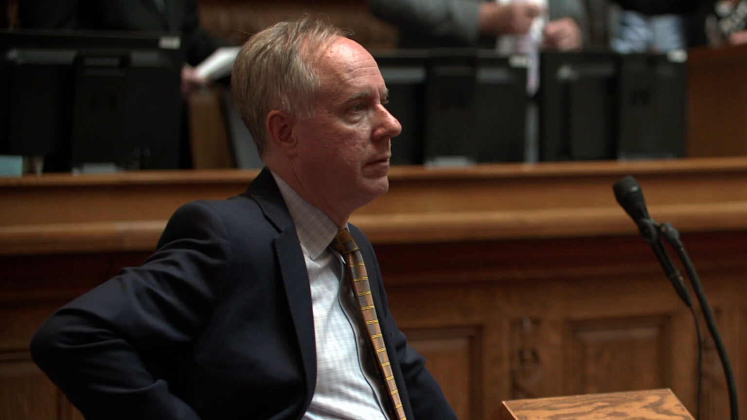 Robin Vos stands behind a wood podium with a microphone and listens, with other people standing at a legislative dais in the background.