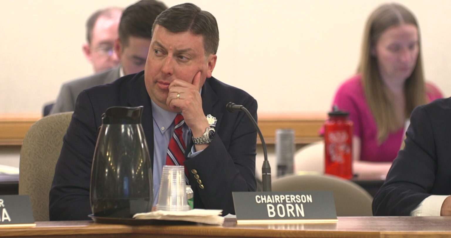 Mark Born holds his left hand to his face while sitting at a table with a microphone, a sign reading Chairperson Born, a carafe and stack of plastic cups, with other people who are out of focus seated in the background.
