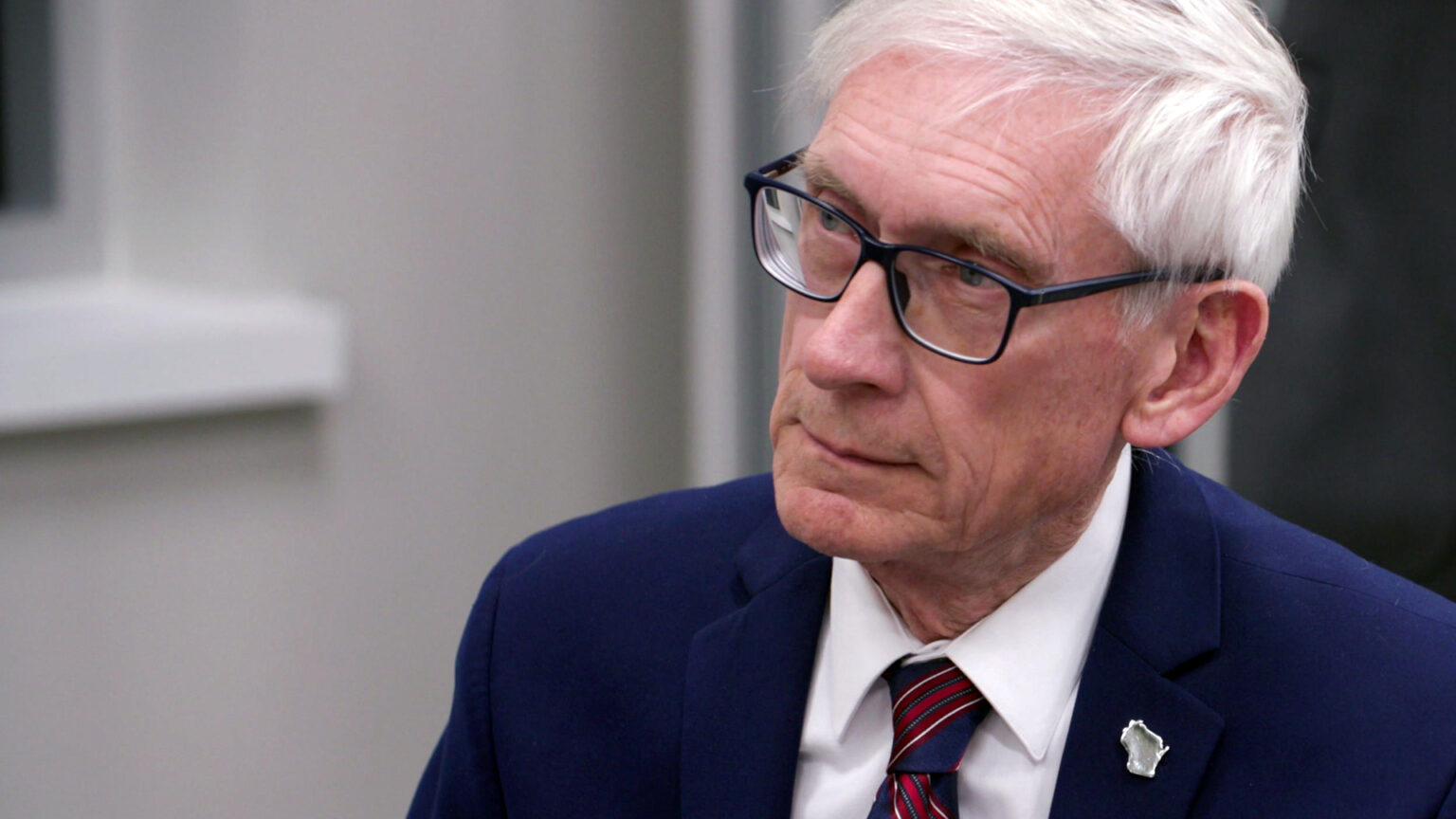Tony Evers sits and listens to another person in a room with a windowsill in the background.
