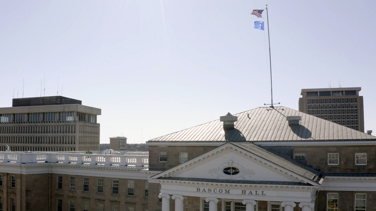 The U.S. and Wisconsin flags fly at the top of a flagpole affixed to the sloped roof of a multi-story stone masonry building with a pediment atop its front entrance with a sign reading Bascom Hall, with multiple other modernist concrete and glass buildings visible in the background under a clear sky.