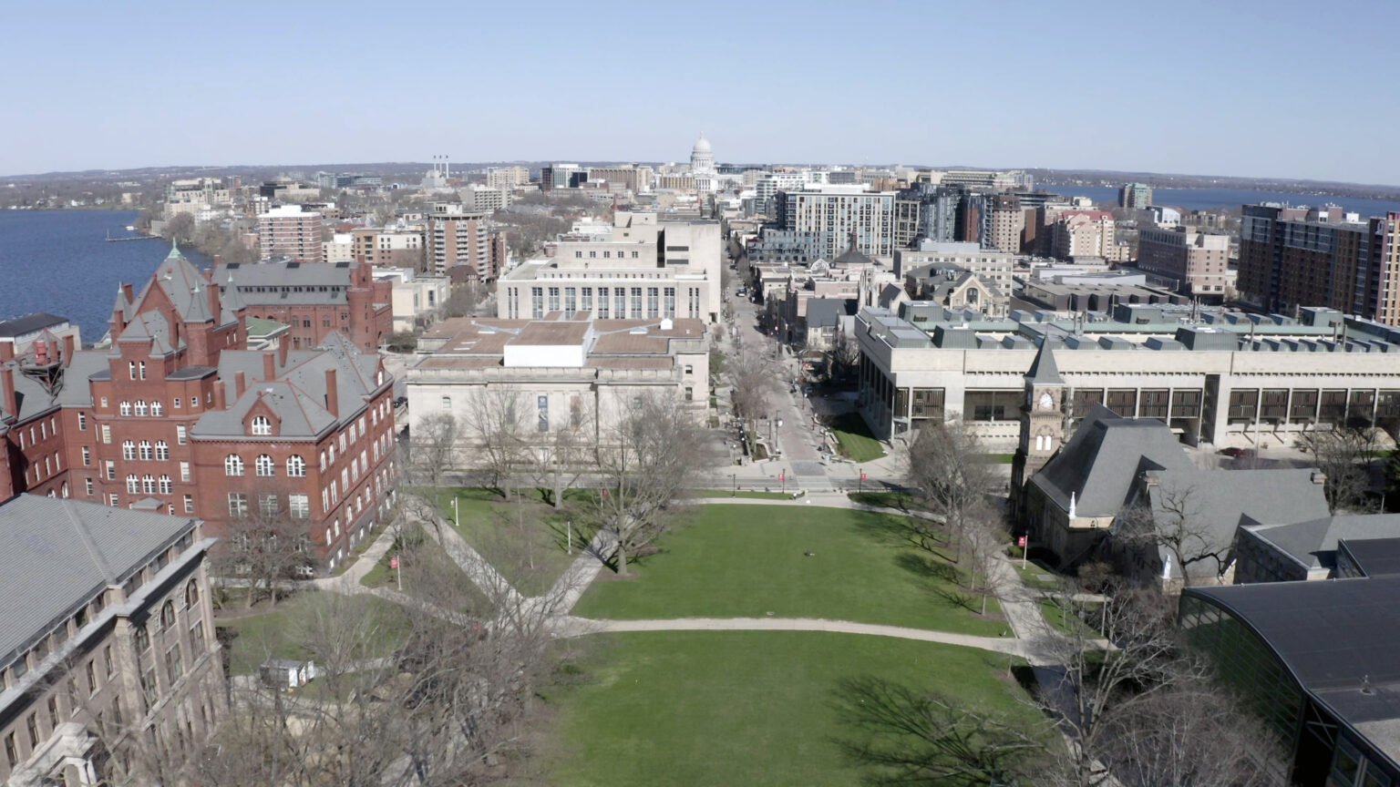 An aerial photo shows areas of grassy lawn at the bottom of a hill, with masonry and concrete buildings on either side, with a cityscape of various buildings, the dome of the Wisconsin State Capitol and portions of two lakes visible in the background.