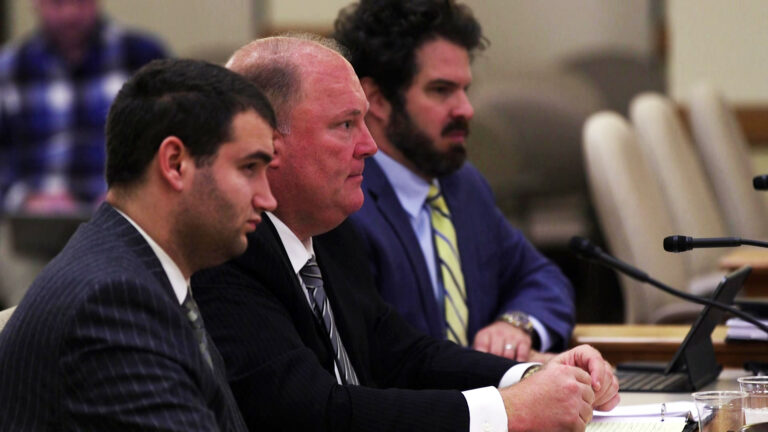 Michael Gableman sits at a table and speaks into a microphone with other people seated on either side of him, in a room with rows of chairs.