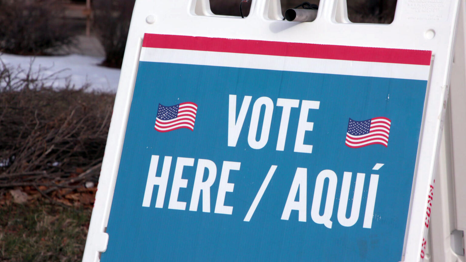 A plastic sandwich board sign with two U.S. flag graphics and the words VOTE HERE / AQUÍ stands next ground covered with fallen leaves and snow.
