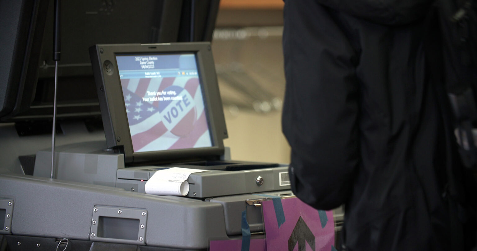 A screen on a ballot tabulator shows a graphic with a U.S. flag and a red-white-and-blue button with the word VOTE, with a piece of paper with an upward pointing arrow taped to its side as a blurry person standing in the foreground approaches the machine.