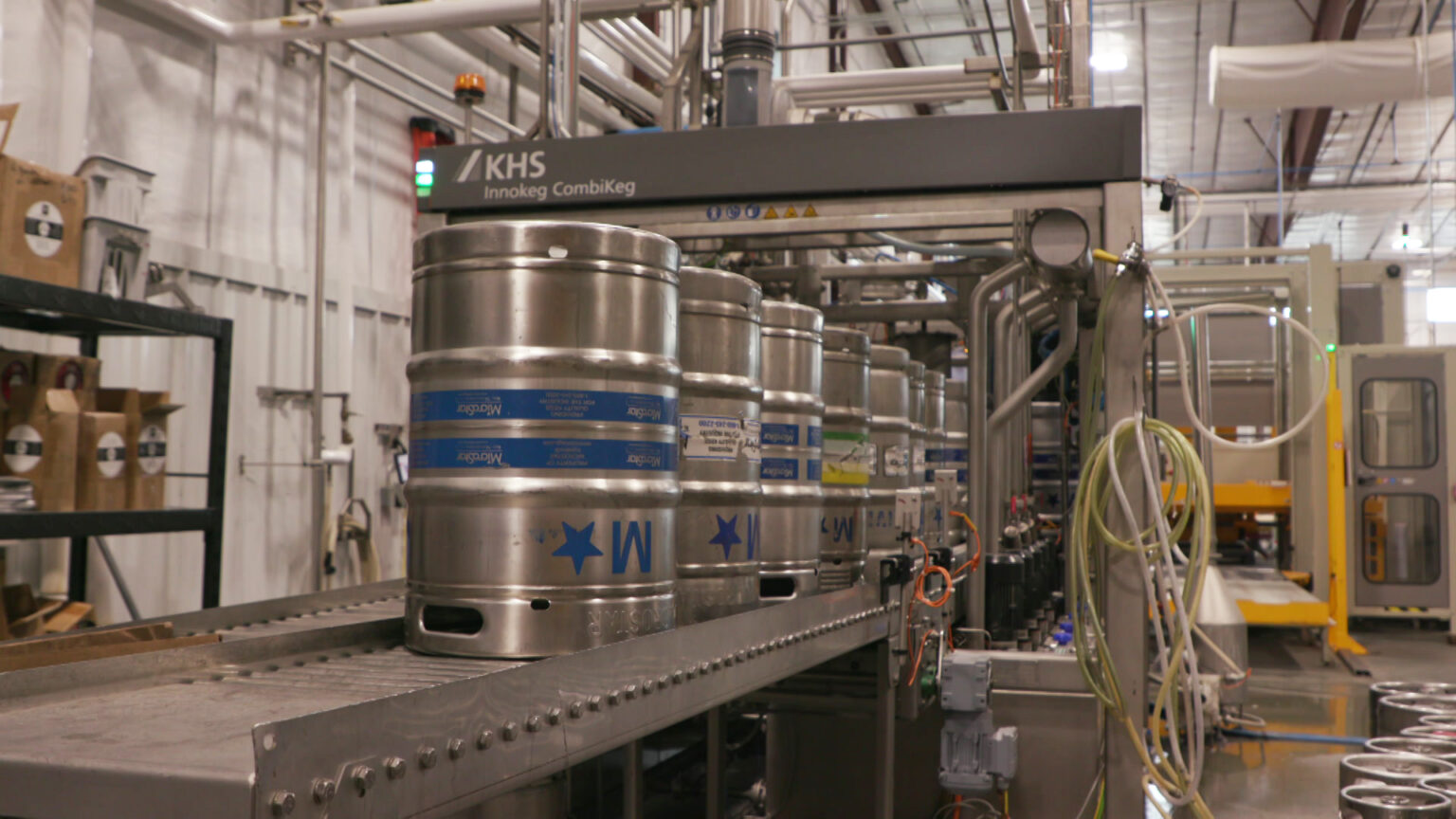 Multiple metal beer kegs move on rollers along a production line in a large room with industrial brewing equipment.