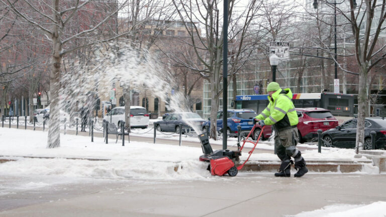 A worker uses a snow blower to remove snow on a walkway, with leafless trees, parked vehicles, a city bus and buildings in the background.