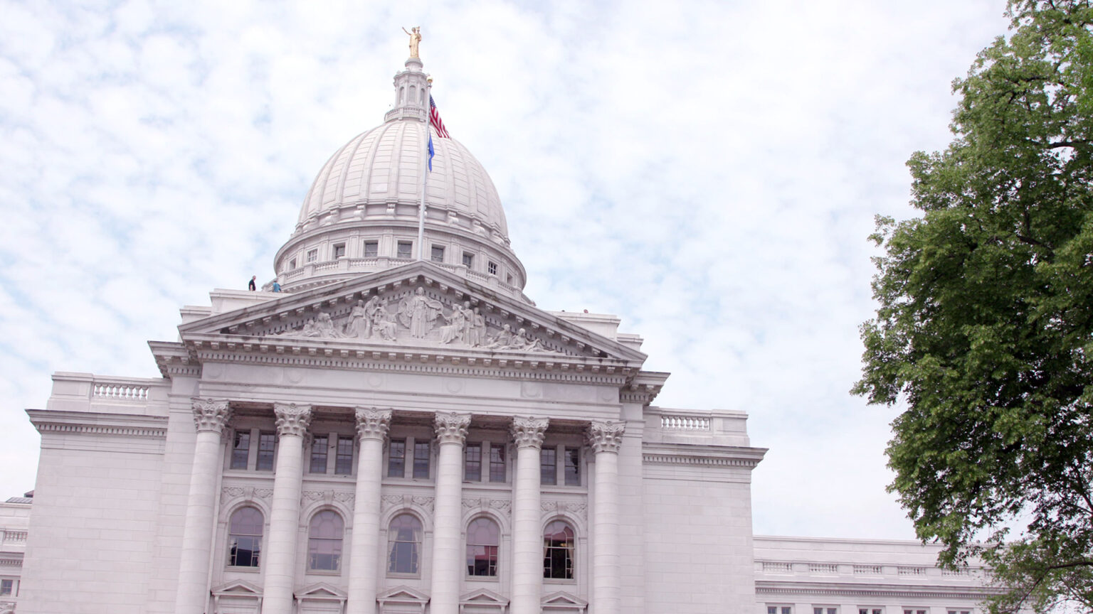 The U.S. and Wisconsin flags fly atop a flagpole on one wing of the Wisconsin Capitol building, framed by its dome in the background and a tree in the foreground.