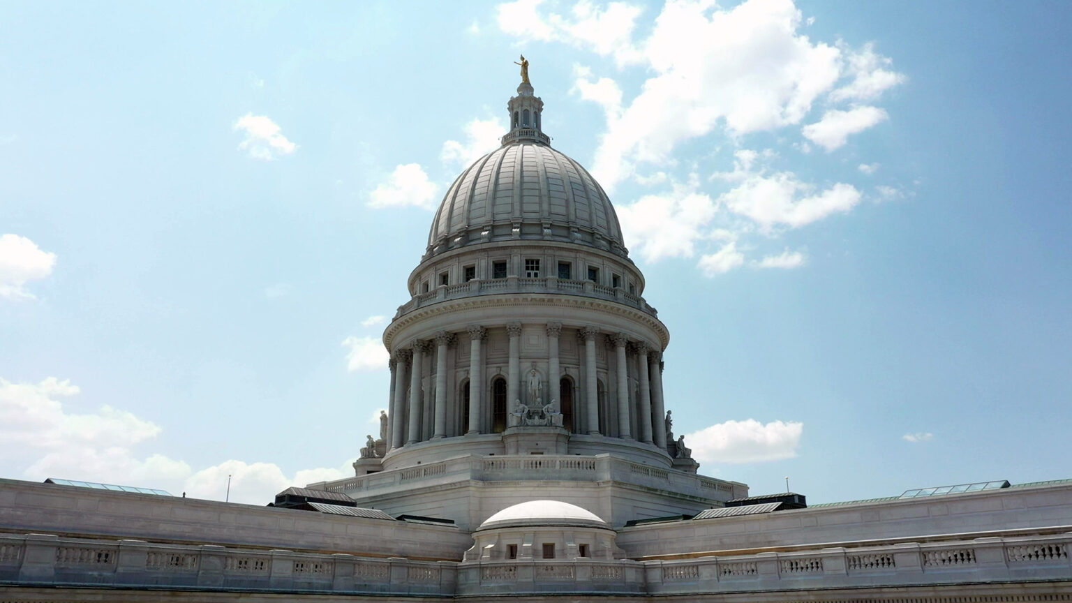 The Wisconsin State Capitol dome and two wings of the building stand under a sky with some clouds.