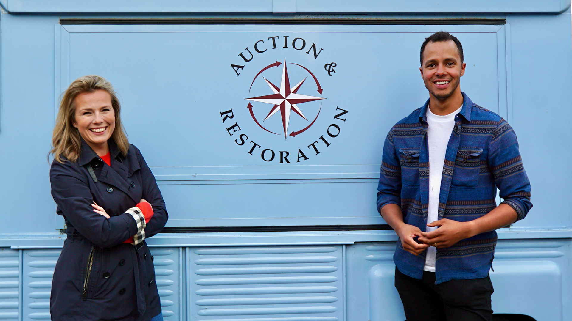 A man and woman stand facing the camera. Both smile. They are in front of a blue van with a logo that reads Auction and Restoration.
