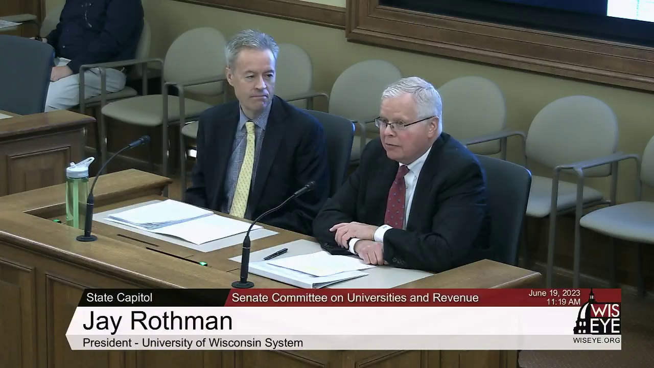 Jay Rothman sits in a chair at a table and speaks into a microphone, with one seated person to his side and another in the background, with a video graphic on the bottom of the image noting a meeting of the Senate Committee on Universities and Revenue.