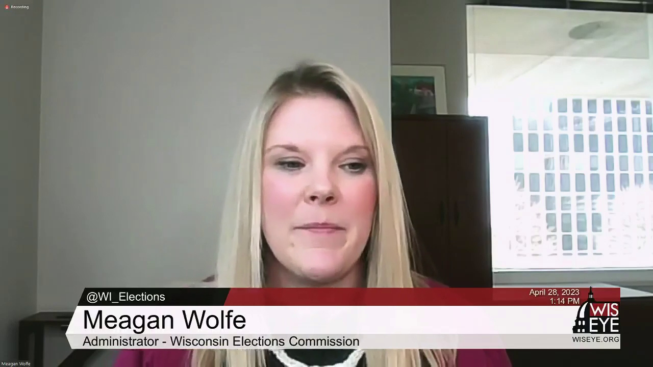 A video still image shows Meagan Wolfe siting in a room with a armoire and window in the background while speaking into a computer camera, with a video graphic on the bottom of the image noting her name and  title as administrator of the Wisconsin Elections Commission.