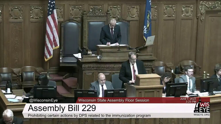 Wisconsin Assembly officers and staff sit and stand at seats on a legislative dais flanked by the U.S. and Wisconsin flags in a room with wood-paneled walls and carvings, with a WisEye-branded video graphic on the bottom of the image noting a Wisconsin State Assembly Floor Session discussion on Assembly Bill 229, described as Prohibiting certain actions by DPS related to the immunization program.