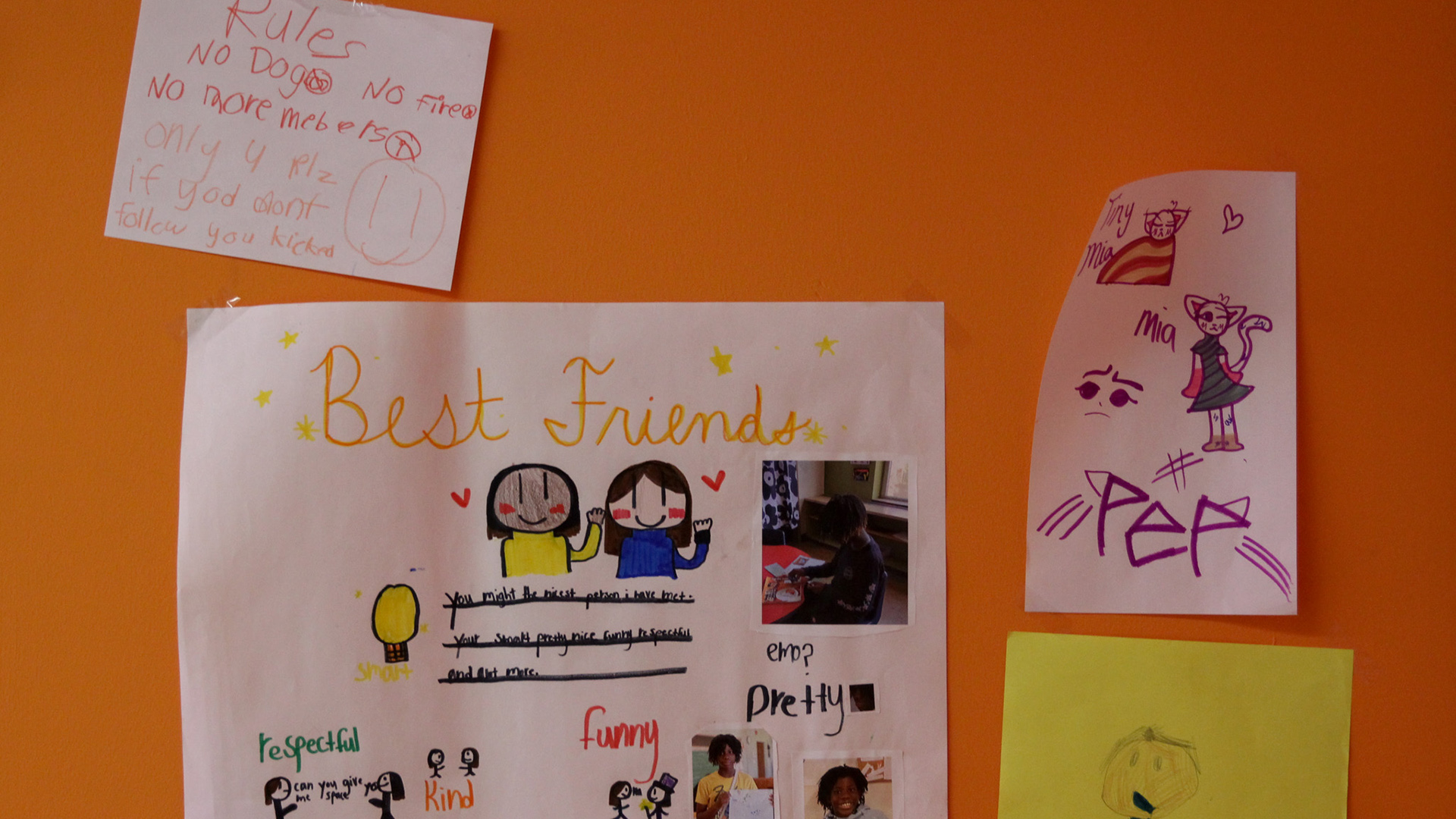 Multiple pieces of paper with children's drawings, writing and photos are taped to a painted wall.