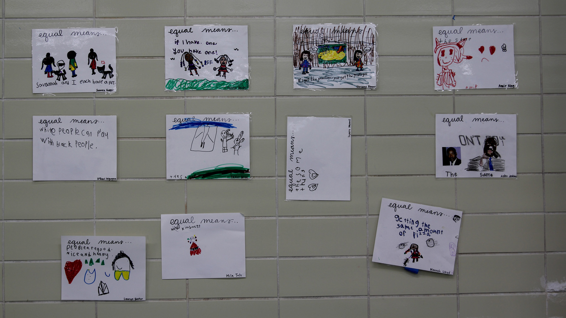 Multiple pieces of paper with children's drawings and writing are taped to a tiled wall.