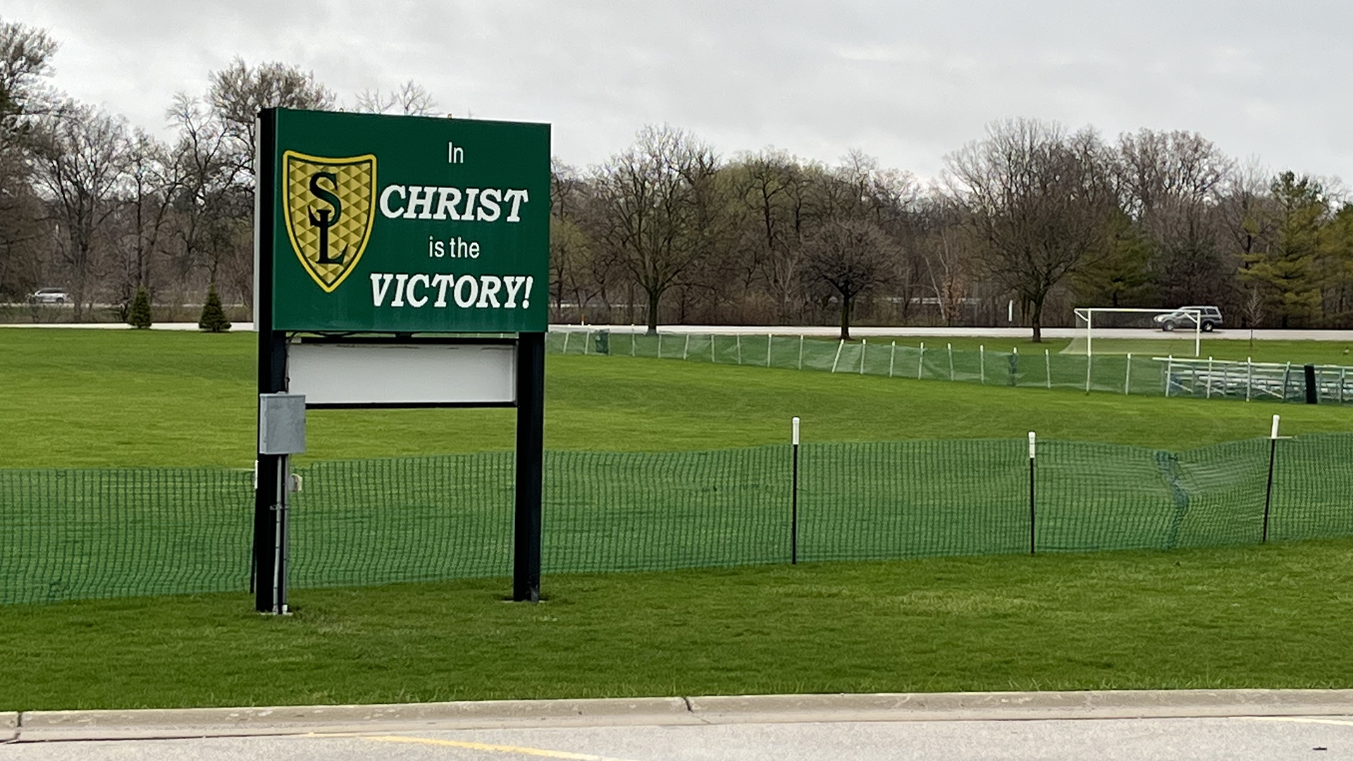 A sign showing a shield logo with the letters "S" and "L" and the words "In Christ Is the Victory!" stands in a lawn surrounded by snow fencing, with a road and line of trees in the background.