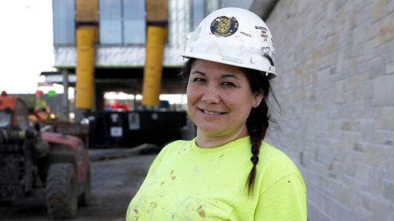 Shannon Pikka poses for a portrait while wearing a safety helmet and standing next to a masonry wall, with two rooftop trash chutes and a construction waste container in the background.