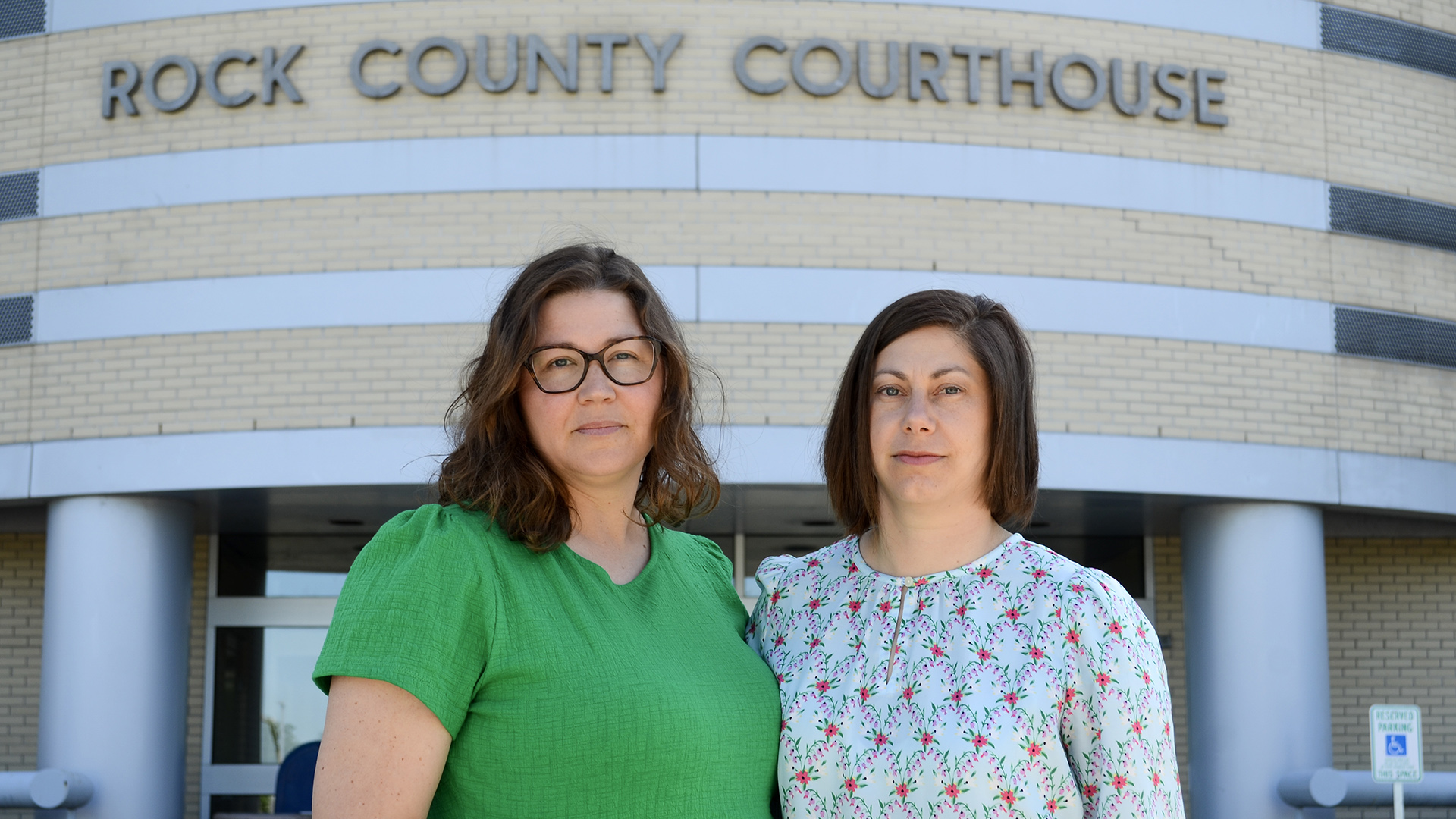 Jamie Gaffke and Ruth Vater stand and pose for a portrait in front of a brick building with metal pillars and a letter sign reading "Rock County Courthouse."