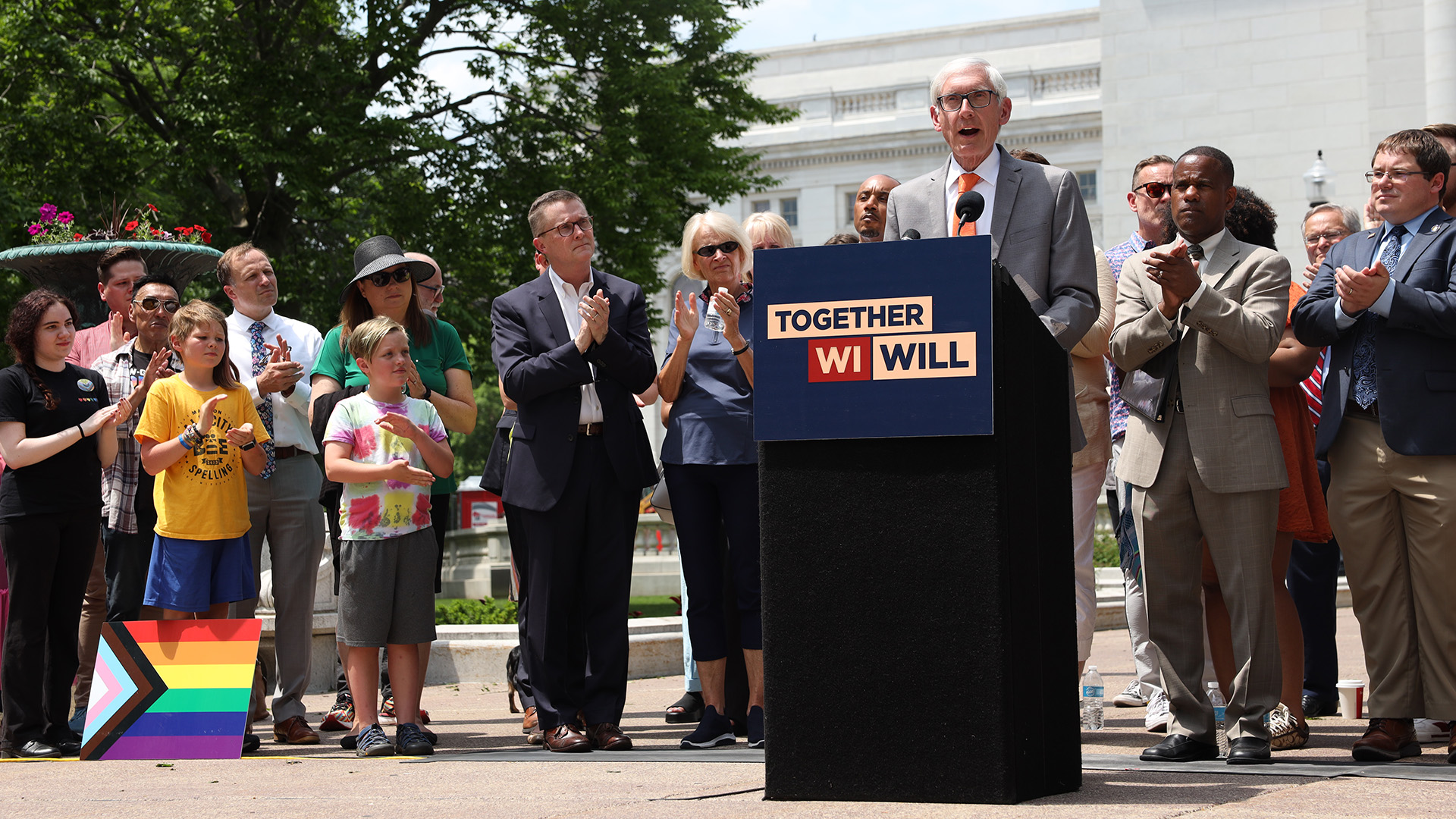 Tony Evers stands behind a cloth-draped podium affixed with a printed sign reading "Together WI Will" and speaks into a microphone as other people standing behind him clap their hands, with a marble masonry building and a tree in the background.