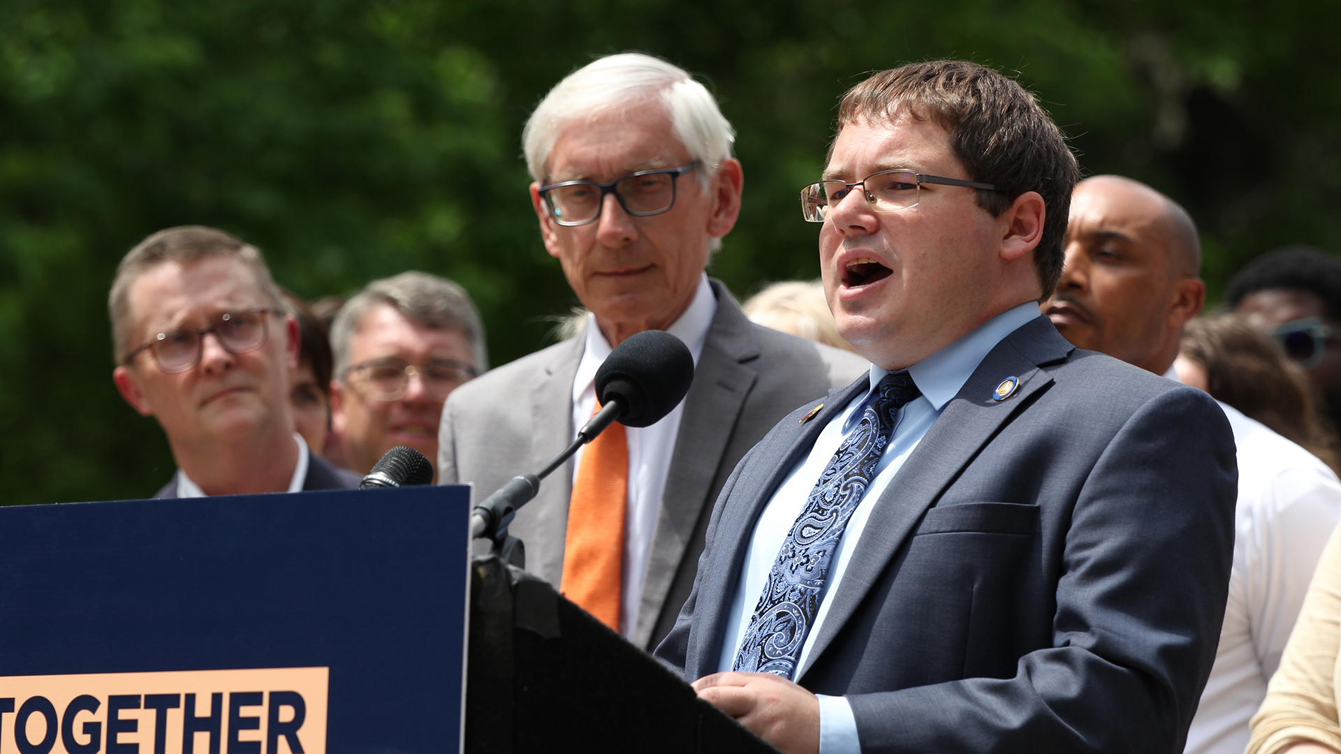 Mark Spreitzer stands behind a podium and speaks into a microphone, with Tony Evers and other people standing in the background.