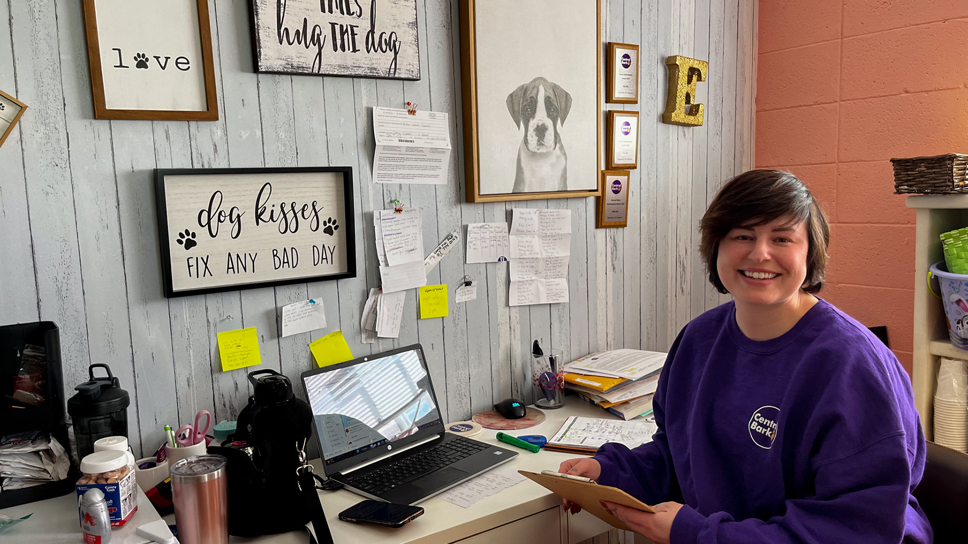 Esther Grams poses for a photo while sitting at a desk with a laptop computer, stacks of paper and other other items, with dog-themed art, award plaques and sheets of paper attached to a painted wood-paneled wall in the background.