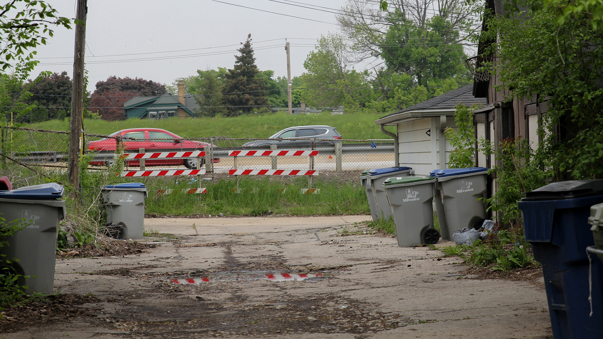 Cars drive on a highway just past the end of an alley lined with trash and recycling receptacles placed outside of residential garages and houses.