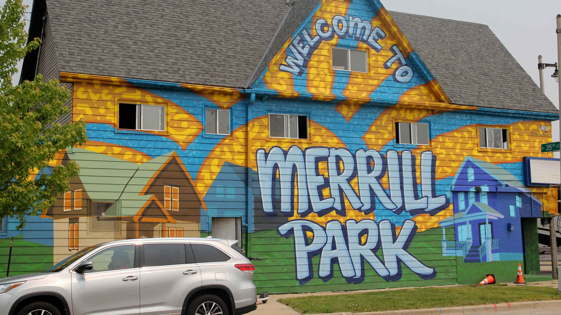 A mural featuring houses, sunrays and the words "Welcome to Merrill Park" is painted on the side of a three-story building, with a parked car in the foreground.
