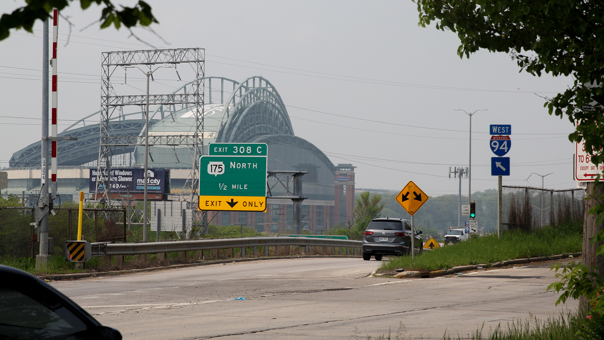 A vehicle drives along the entrance to a highway, with trees in the foreground and the Milwaukee Brewers stadium visible in the background.